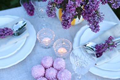Photo of Beautiful lilac flowers, zephyrs, plates, cutlery, glasses and burning candles on table in garden, above view