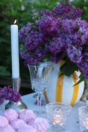 Photo of Beautiful lilac flowers in vase, zephyrs, glasses and burning candles on table in garden, closeup