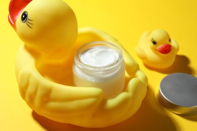 Photo of Jar of moisturizing body cream and rubber ducks on yellow background, closeup