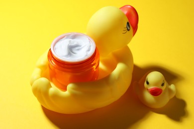 Photo of Jar of moisturizing body cream and rubber ducks on yellow background, closeup