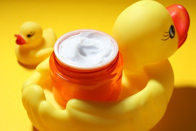 Photo of Jar of moisturizing body cream and rubber ducks on yellow background, closeup