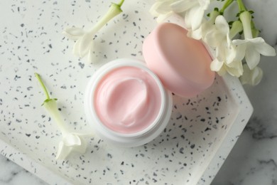 Photo of Jar of moisturizing body cream and flowers on white marble table, flat lay