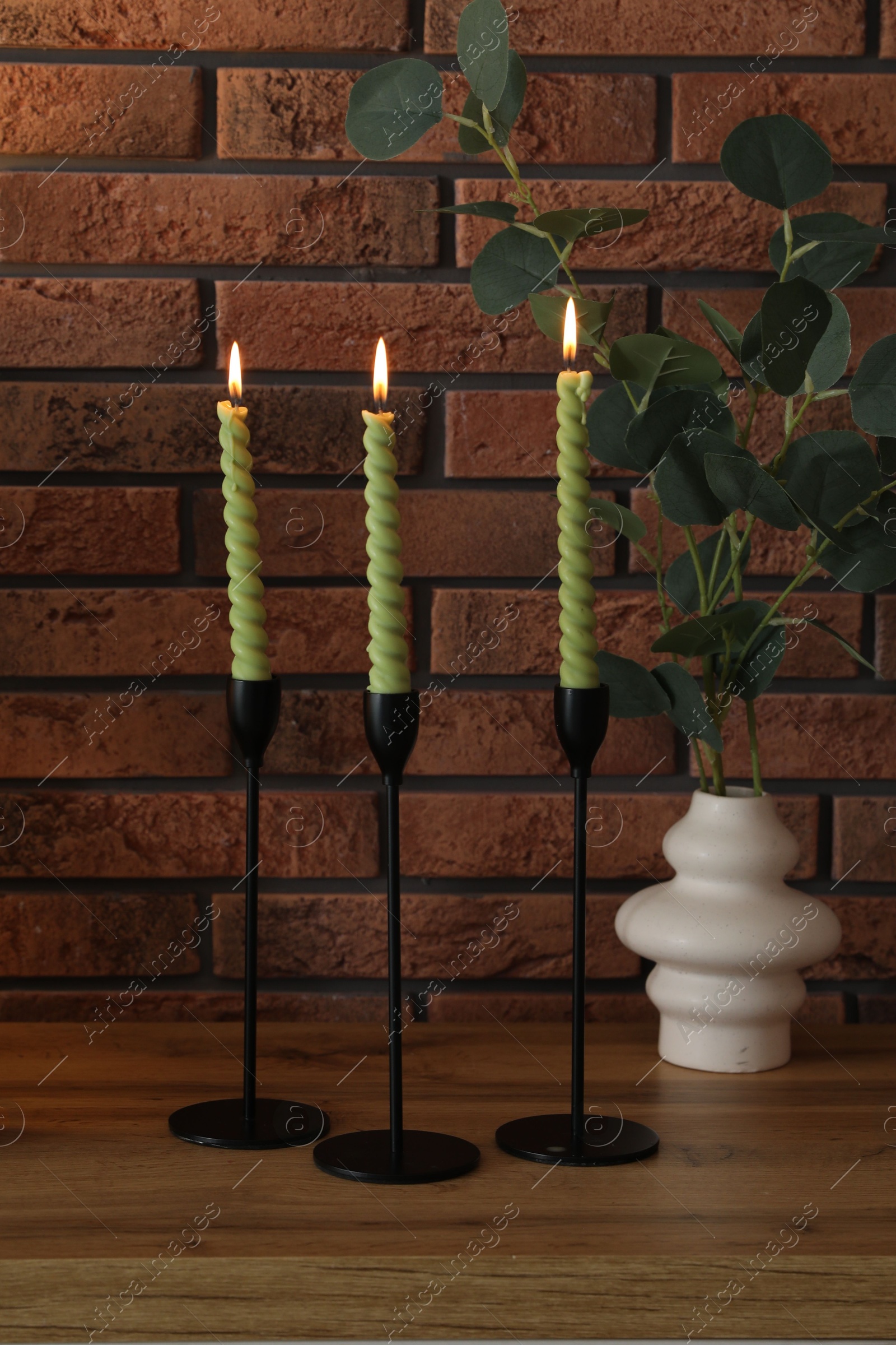 Candlesticks with burning candles on table near brick wall Photo of Candlesticks with burning candles on table near brick wall