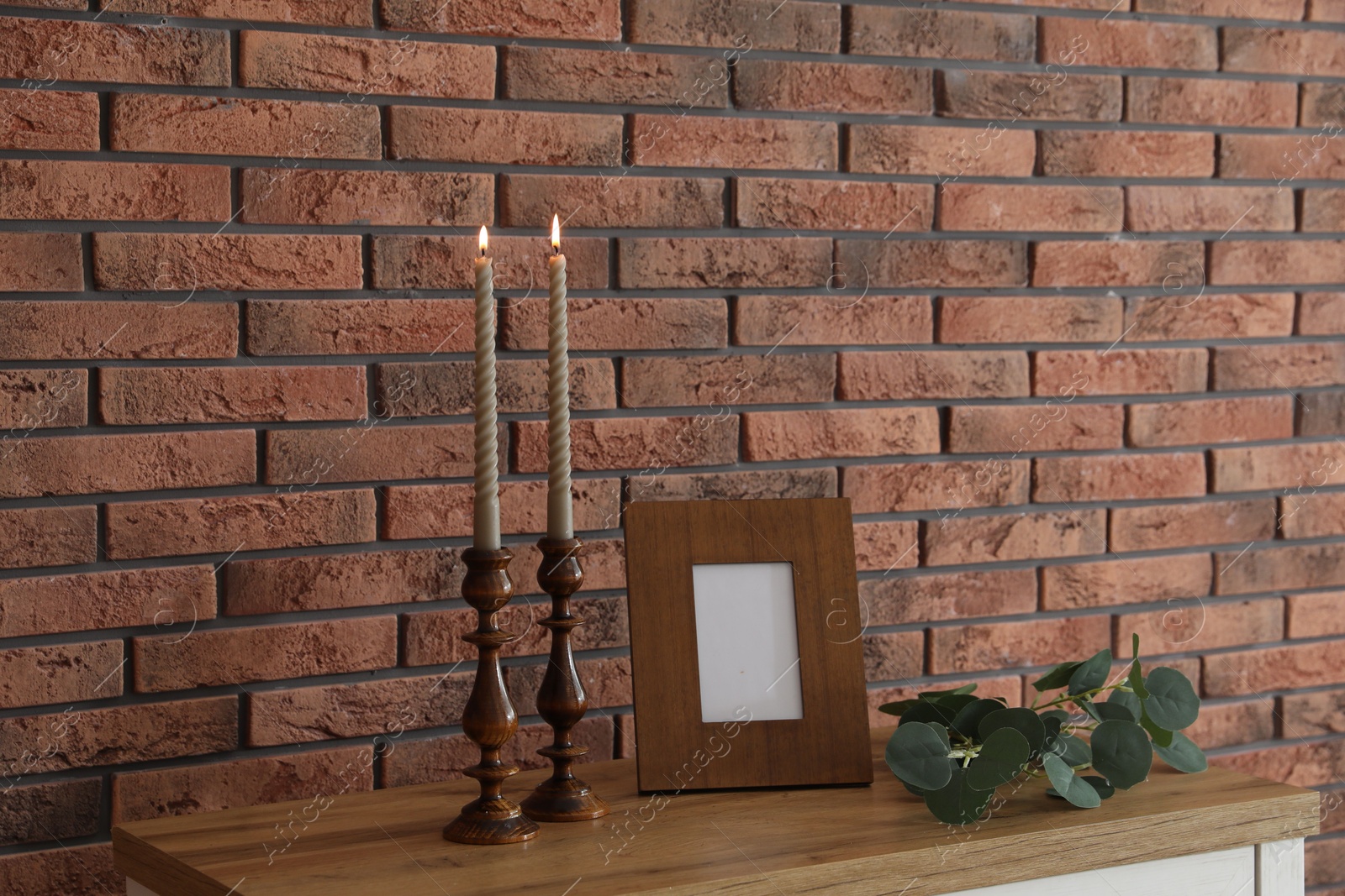 Candlesticks with burning candles on table near brick wall Photo of Candlesticks with burning candles on table near brick wall