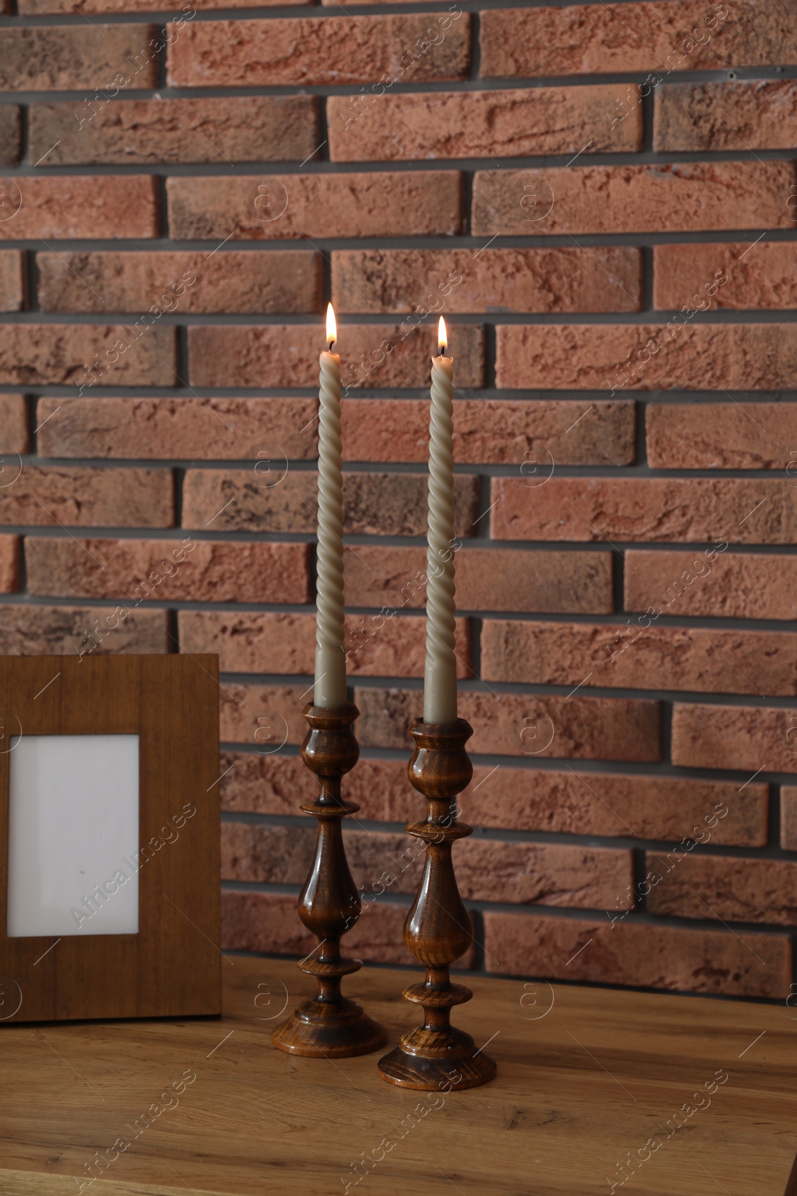 Candlesticks with burning candles on table near brick wall Photo of Candlesticks with burning candles on table near brick wall