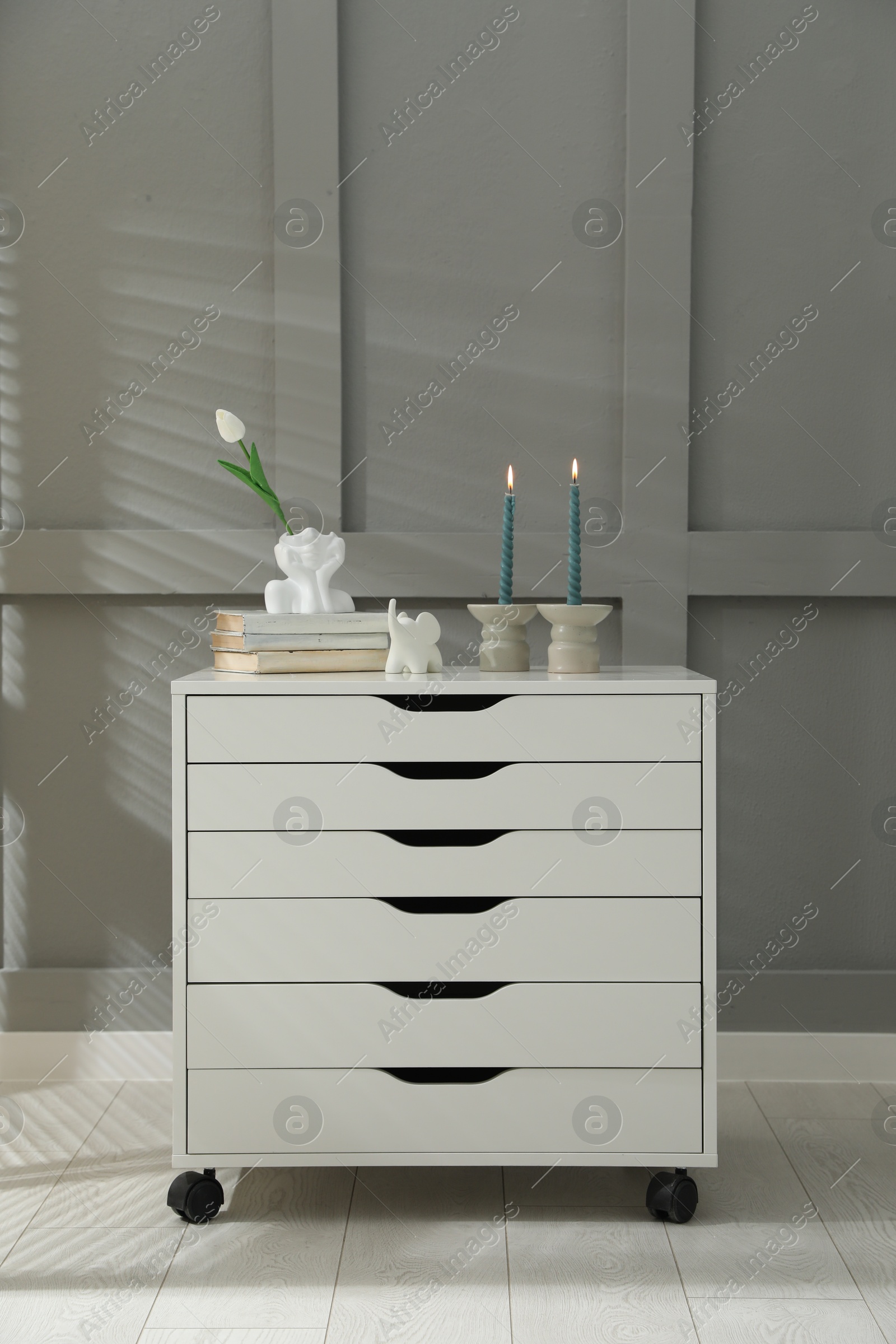 Candlesticks with burning candles on chest of drawers near grey wall Photo of Candlesticks with burning candles on chest of drawers near grey wall