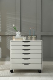 Photo of Candlesticks with burning candles on chest of drawers near grey wall