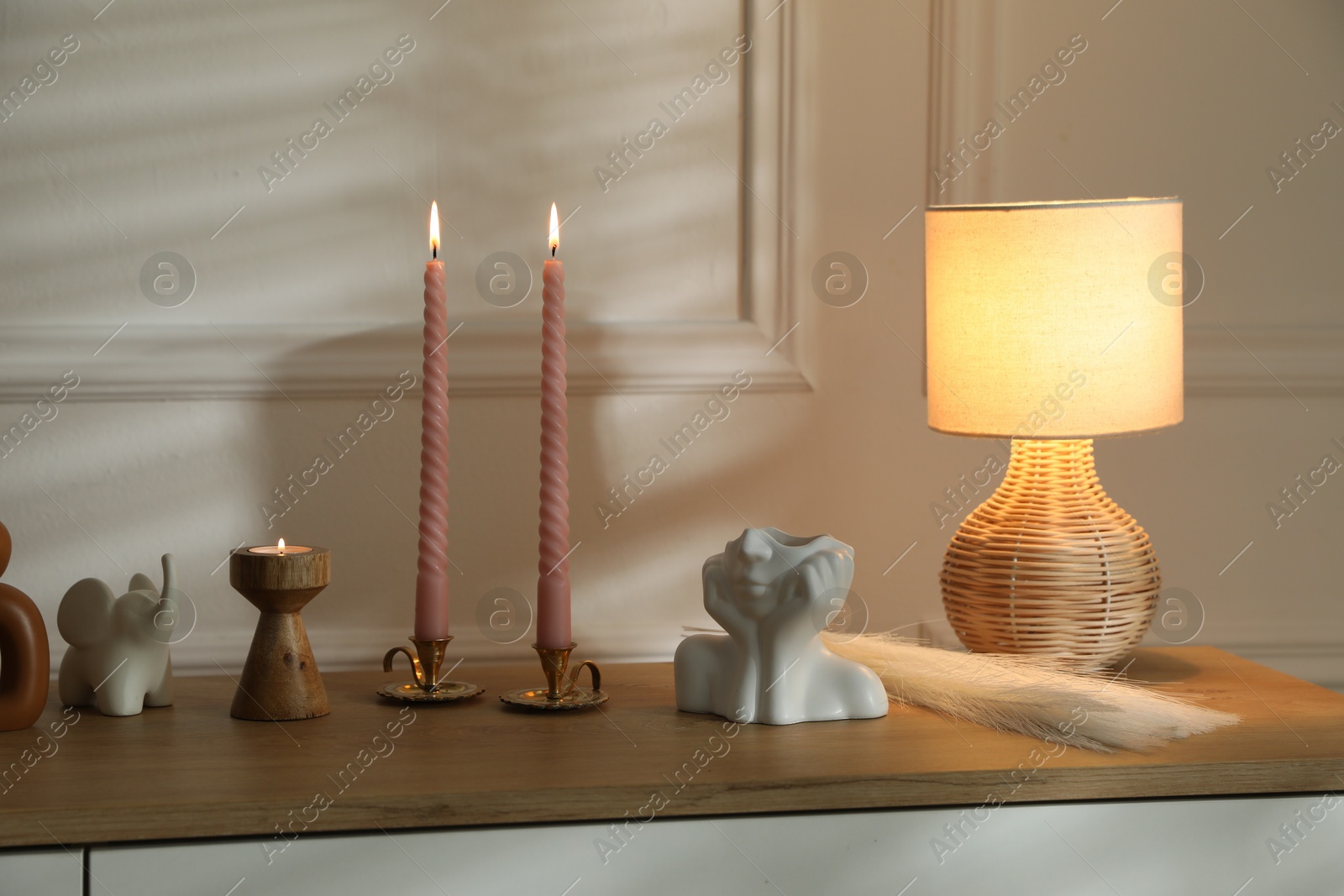 Candlesticks with burning candles on chest of drawers near white wall Photo of Candlesticks with burning candles on chest of drawers near white wall