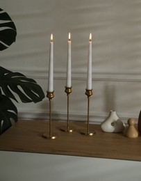 Photo of Candlesticks with burning candles on chest of drawers near white wall