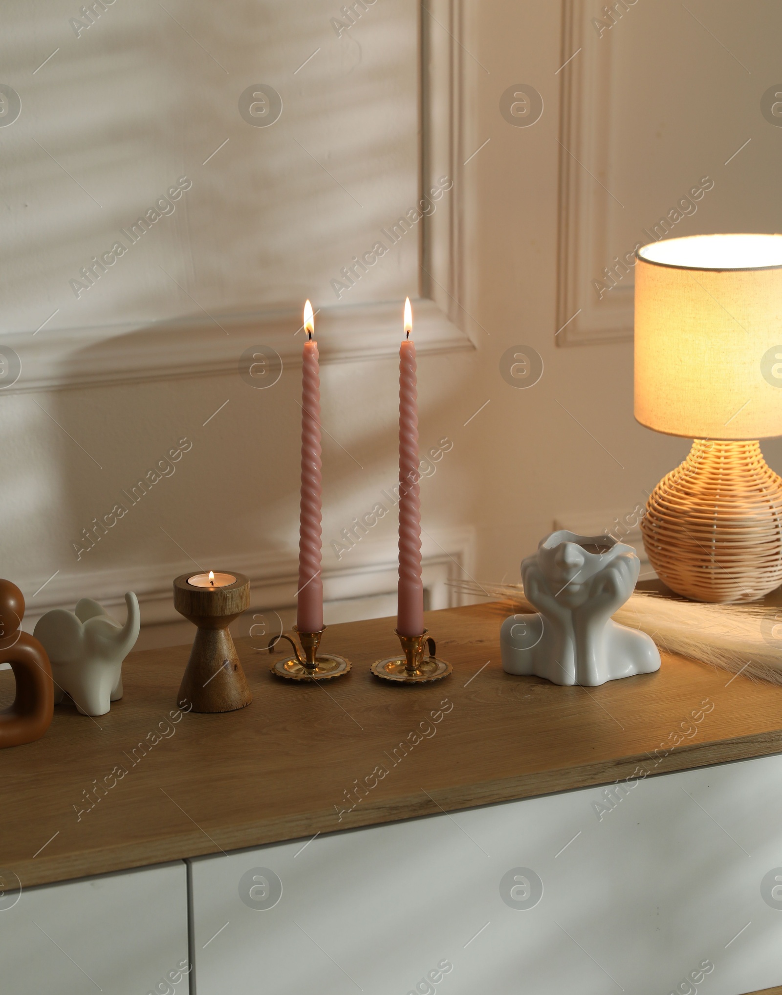 Candlesticks with burning candles on chest of drawers near white wall Photo of Candlesticks with burning candles on chest of drawers near white wall