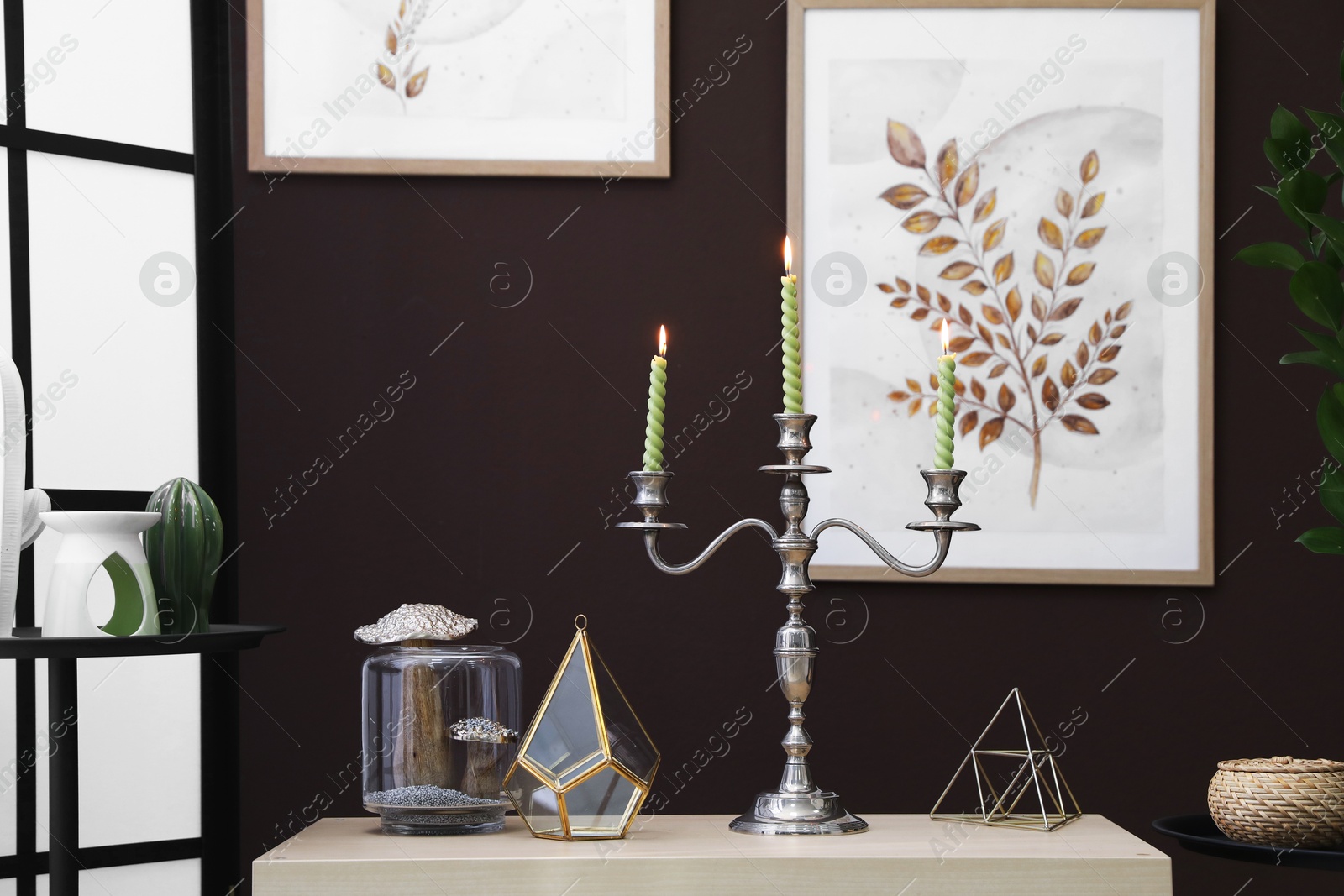 Elegant candlestick with burning candles and decor on cabinet near brown wall Photo of Elegant candlestick with burning candles and decor on cabinet near brown wall