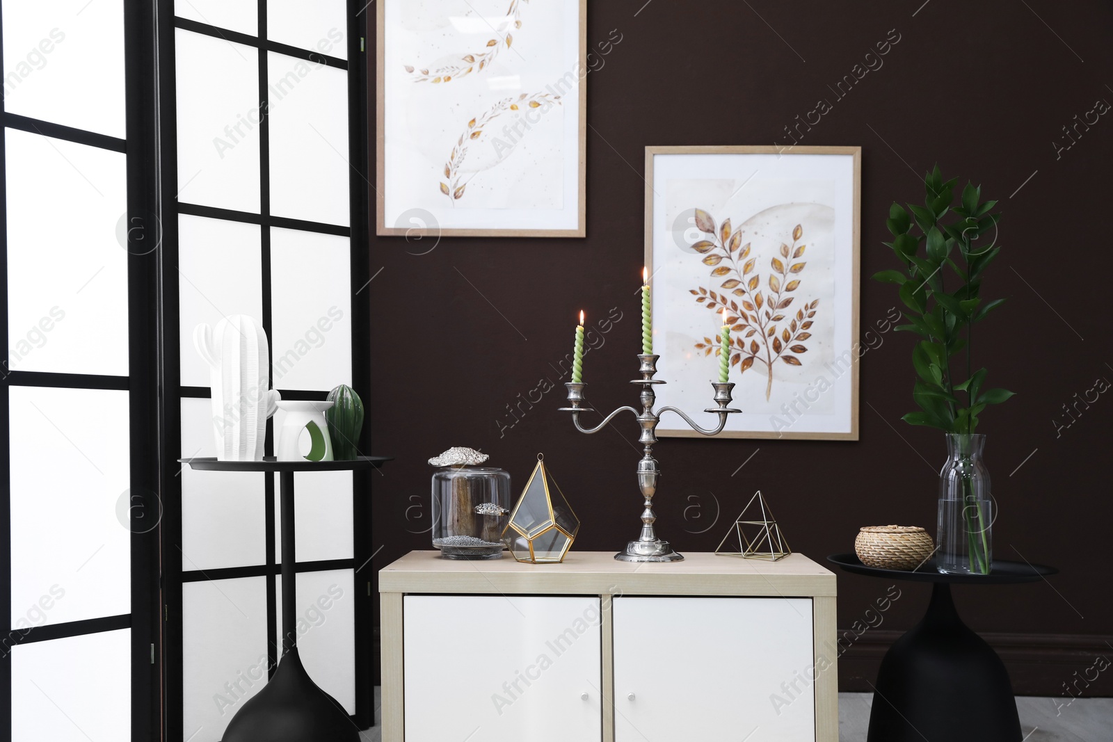 Elegant candlestick with burning candles and decor on cabinet near brown wall Photo of Elegant candlestick with burning candles and decor on cabinet near brown wall