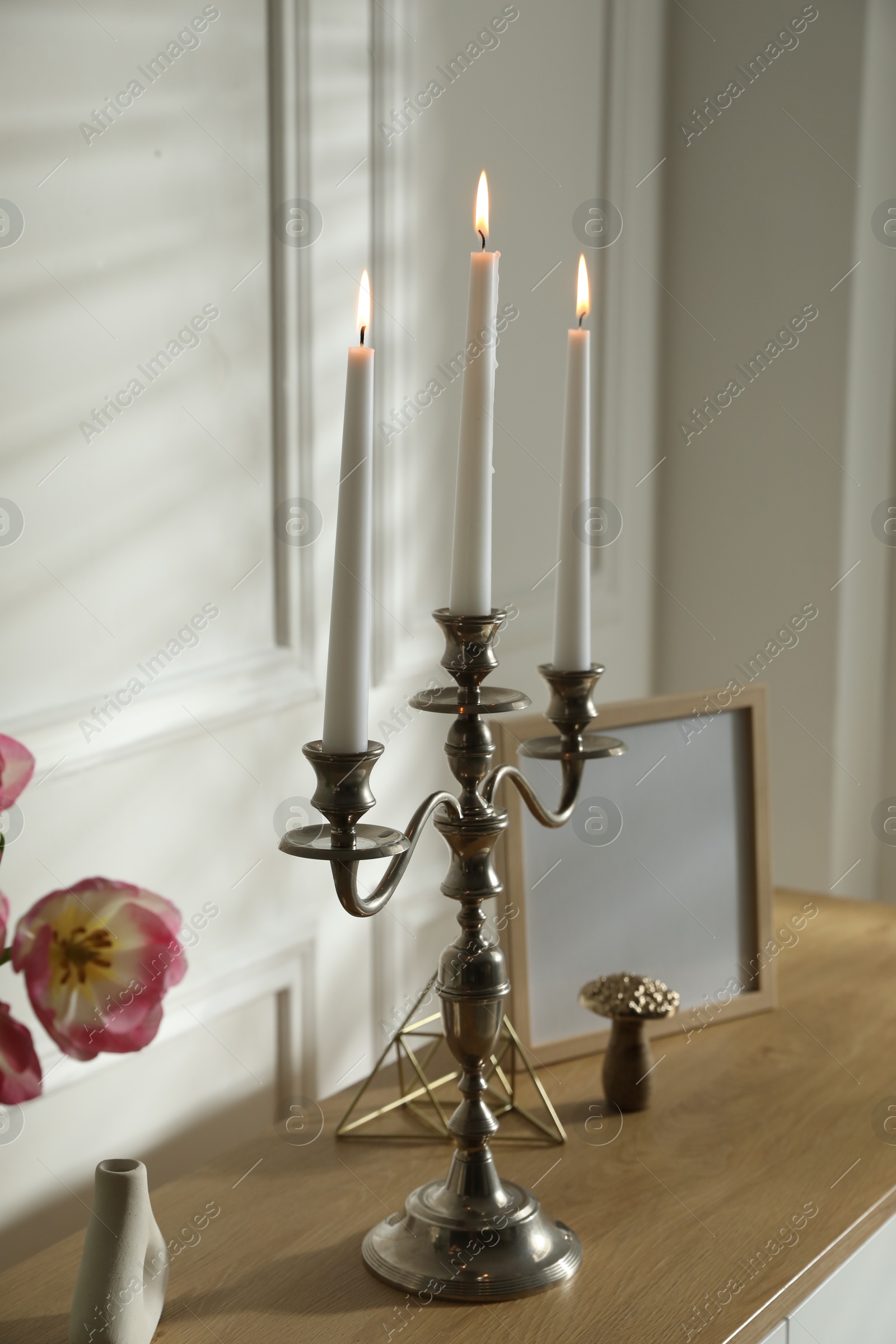 Elegant candlestick with burning candles and decor on table near white wall Photo of Elegant candlestick with burning candles and decor on table near white wall