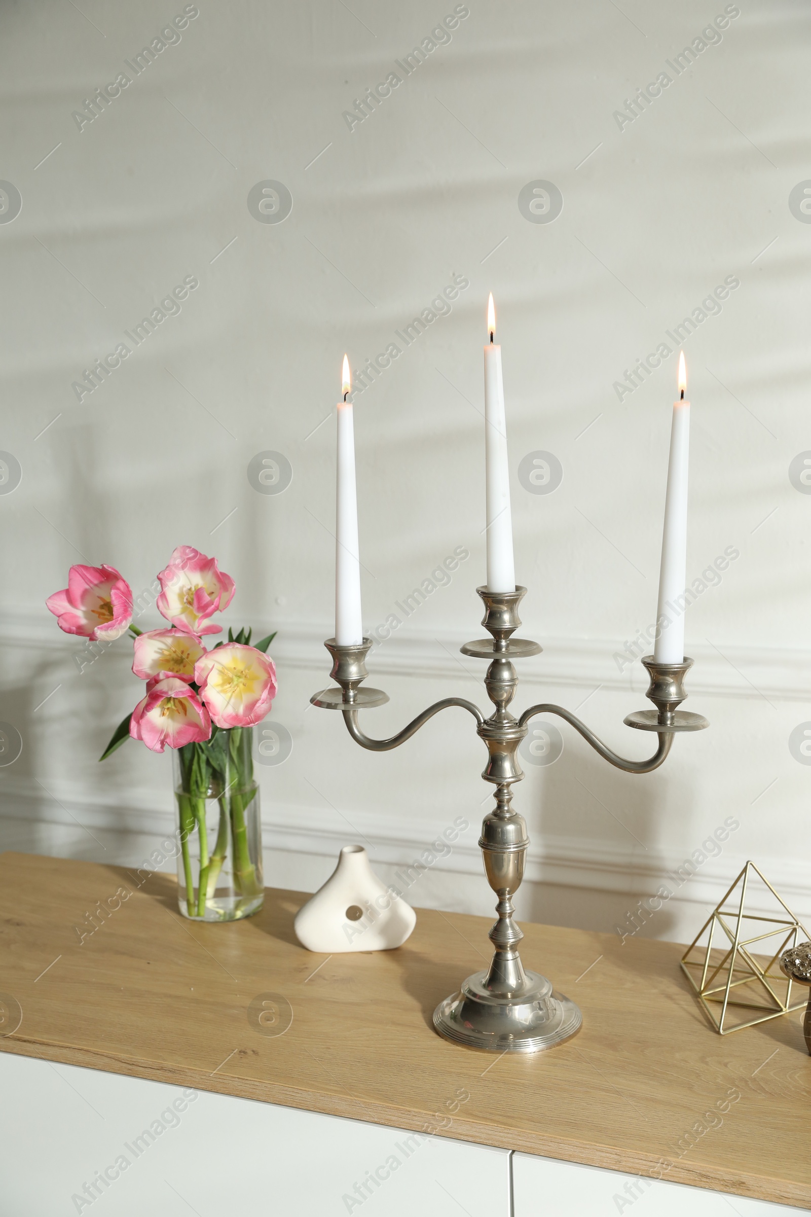 Elegant candlestick with burning candles and decor on chest of drawers near white wall Photo of Elegant candlestick with burning candles and decor on chest of drawers near white wall
