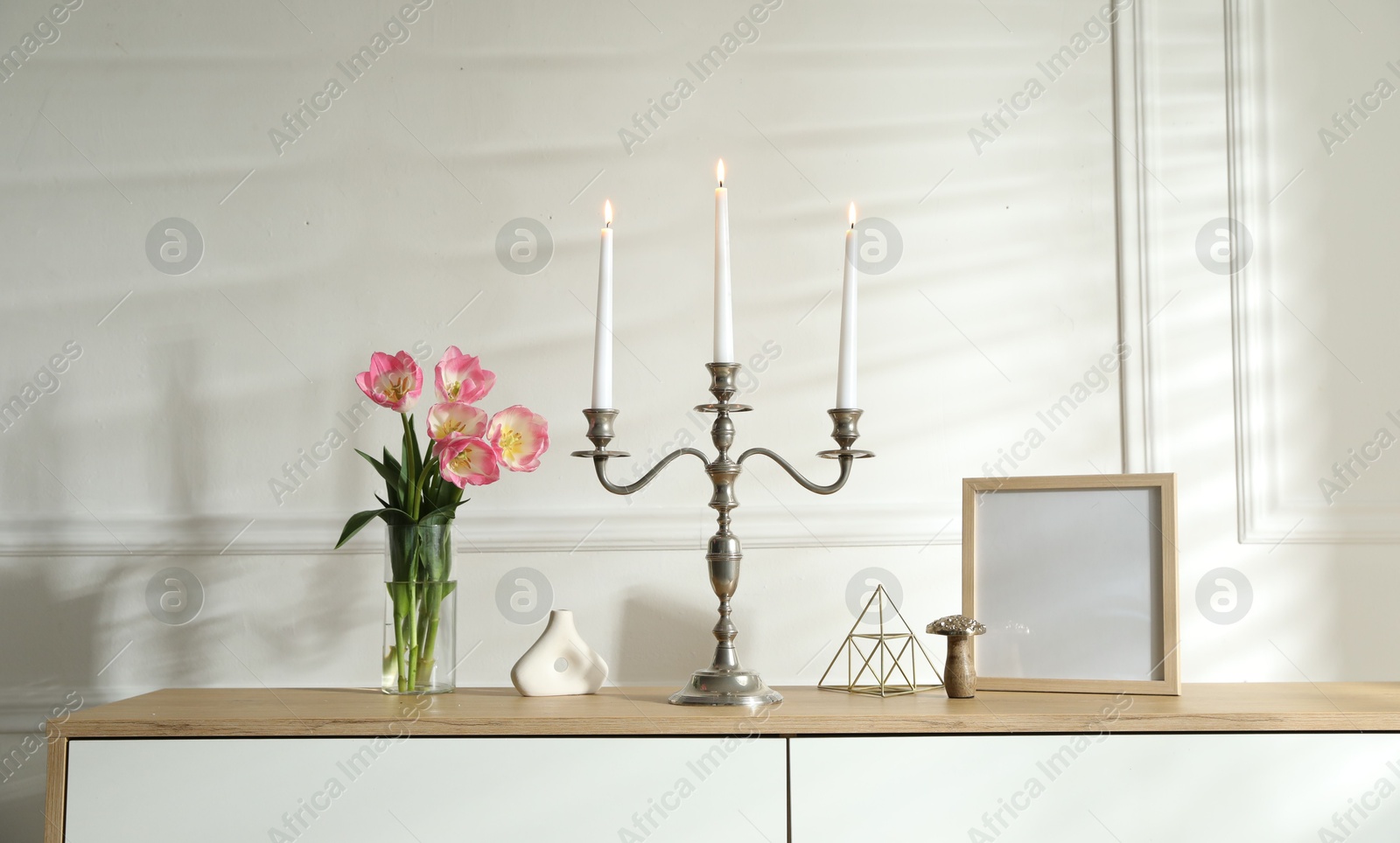 Elegant candlestick with burning candles and decor on chest of drawers near white wall Photo of Elegant candlestick with burning candles and decor on chest of drawers near white wall