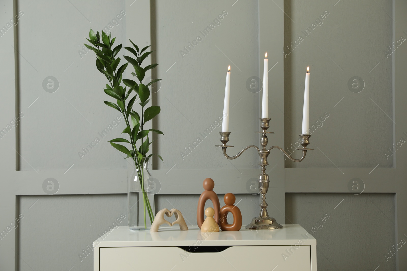 Elegant candlestick with burning candles and decor on chest of drawers near light wall Photo of Elegant candlestick with burning candles and decor on chest of drawers near light wall