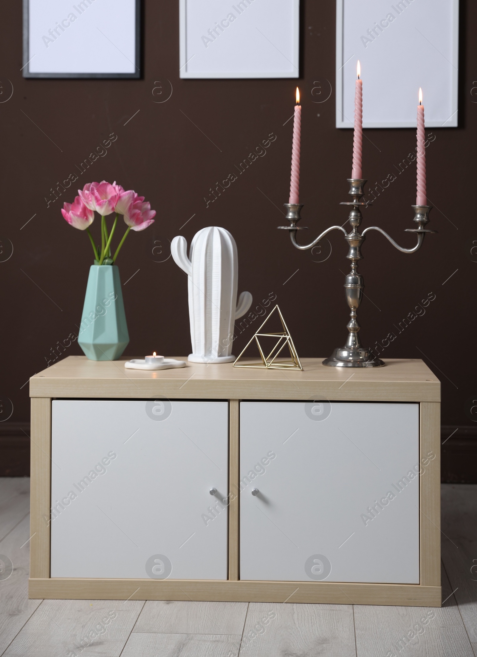 Elegant candlestick with burning candles and decor on cabinet near brown wall Photo of Elegant candlestick with burning candles and decor on cabinet near brown wall