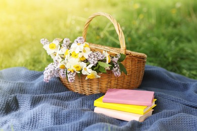 Photo of Stacked books, blanket and wicker basket with flowers on green grass outdoors