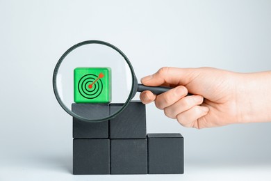 Image of Targeting. Woman holding magnifying glass near green cube with bullseye on light background, closeup
