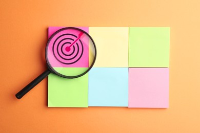 Image of Targeting. Paper notes with bullseye and magnifying glass on orange background, top view