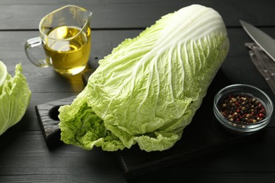 Photo of Fresh Chinese cabbages, oil, knife and peppercorns on black wooden table, closeup