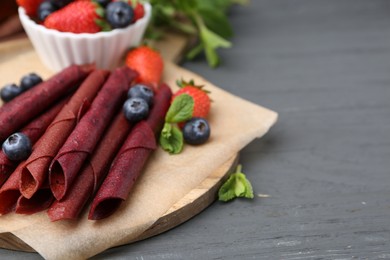 Photo of Tasty fruit leather rolls, strawberries, blueberries and mint on grey wooden table, closeup. Space for text
