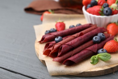 Photo of Tasty fruit leather rolls, strawberries, blueberries and mint on grey wooden table, closeup. Space for text