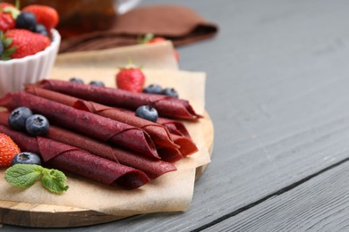 Photo of Tasty fruit leather rolls, strawberries, blueberries and mint on grey wooden table, closeup. Space for text
