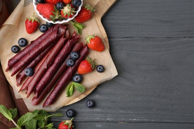 Photo of Tasty fruit leather rolls, strawberries, blueberries and mint on grey wooden table, flat lay. Space for text