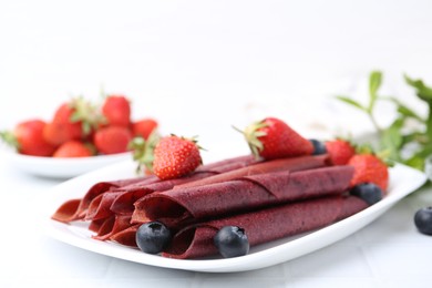 Photo of Tasty fruit leather rolls, strawberries and blueberries on white table, closeup. Space for text