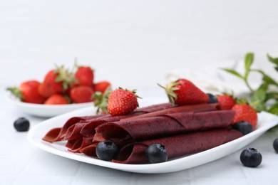 Photo of Tasty fruit leather rolls, strawberries and blueberries on white table, closeup. Space for text