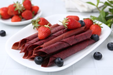 Photo of Tasty fruit leather rolls, strawberries and blueberries on white table, closeup