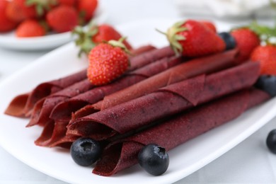 Photo of Tasty fruit leather rolls, strawberries and blueberries on white table, closeup