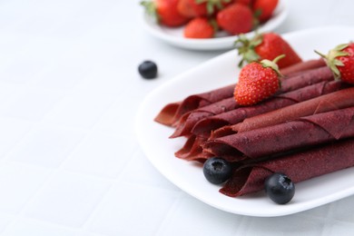 Photo of Tasty fruit leather rolls, strawberries and blueberries on white table, closeup. Space for text