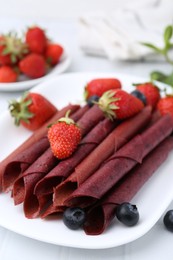 Photo of Tasty fruit leather rolls, strawberries and blueberries on white table, closeup