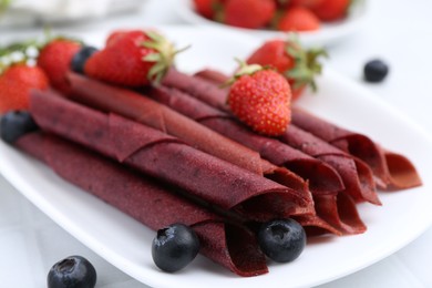 Photo of Tasty fruit leather rolls, strawberries and blueberries on white table, closeup