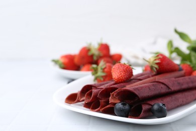 Photo of Tasty fruit leather rolls, strawberries and blueberries on white table, closeup. Space for text