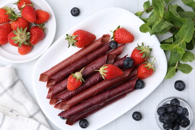 Photo of Tasty fruit leather rolls, strawberries, blueberries and mint on white tiled table, flat lay