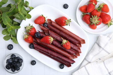 Photo of Tasty fruit leather rolls, strawberries, blueberries and mint on white tiled table, flat lay