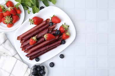 Photo of Tasty fruit leather rolls, strawberries, blueberries and mint on white tiled table, flat lay. Space for text