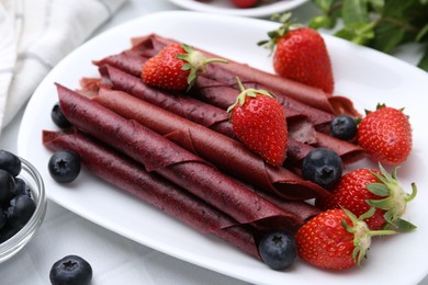 Photo of Tasty fruit leather rolls, strawberries and blueberries on white table, closeup