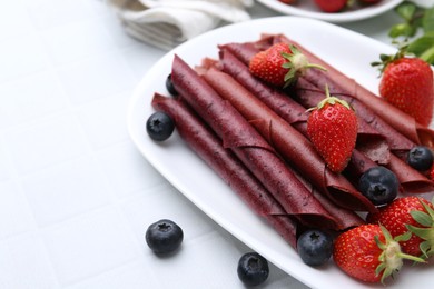 Photo of Tasty fruit leather rolls, strawberries and blueberries on white table, closeup. Space for text