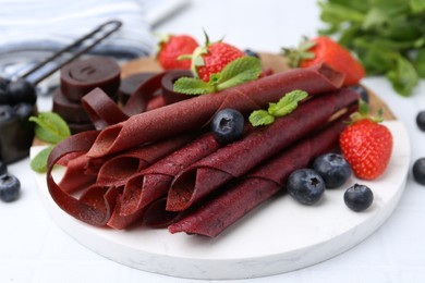 Photo of Tasty fruit leather rolls, strawberries, blueberries and mint on white table, closeup