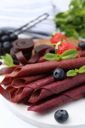 Photo of Tasty fruit leather rolls, strawberries, blueberries and mint on white table, closeup