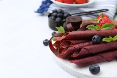 Photo of Tasty fruit leather rolls, strawberries, blueberries and mint on white tiled table, closeup. Space for text