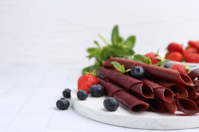 Photo of Tasty fruit leather rolls, strawberries, blueberries and mint on white tiled table, closeup. Space for text