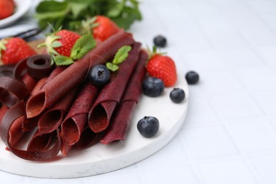 Photo of Tasty fruit leather rolls, strawberries, blueberries and mint on white tiled table, closeup