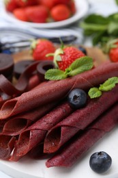 Photo of Tasty fruit leather rolls, strawberries, blueberries and mint on white table, closeup
