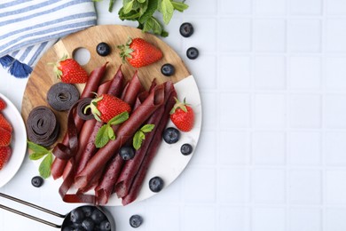 Photo of Tasty fruit leather rolls, strawberries, blueberries and mint on white tiled table, flat lay. Space for text