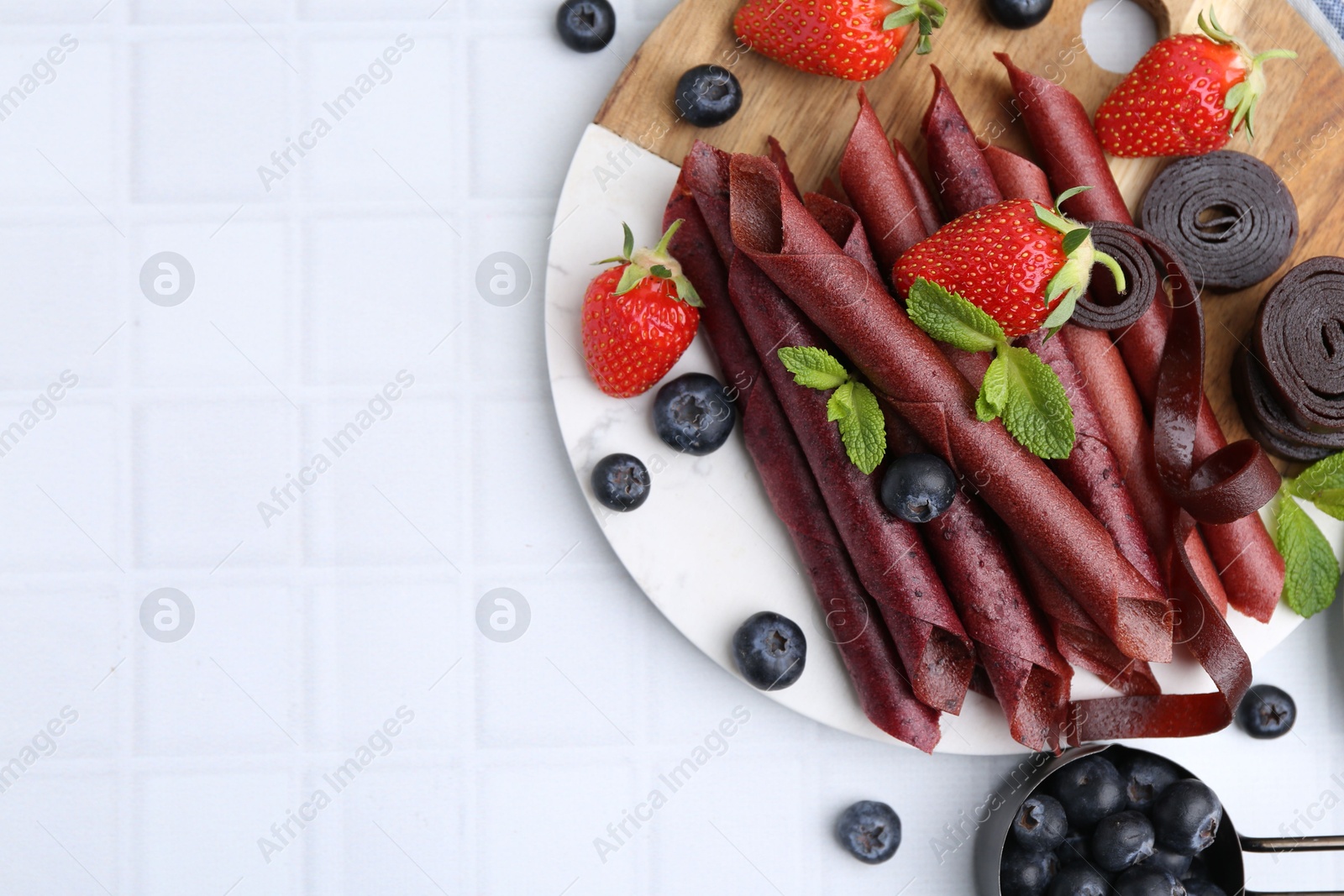 Photo of Tasty fruit leather rolls, strawberries, blueberries and mint on white tiled table, flat lay. Space for text